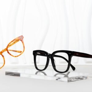 Elegant glass frames in black and tortoise colors showcased on a clear acrylic stand against a wavy backdrop.