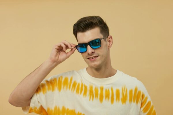 Young man wearing blue sunglasses and a tie-dye shirt poses against a yellow background.